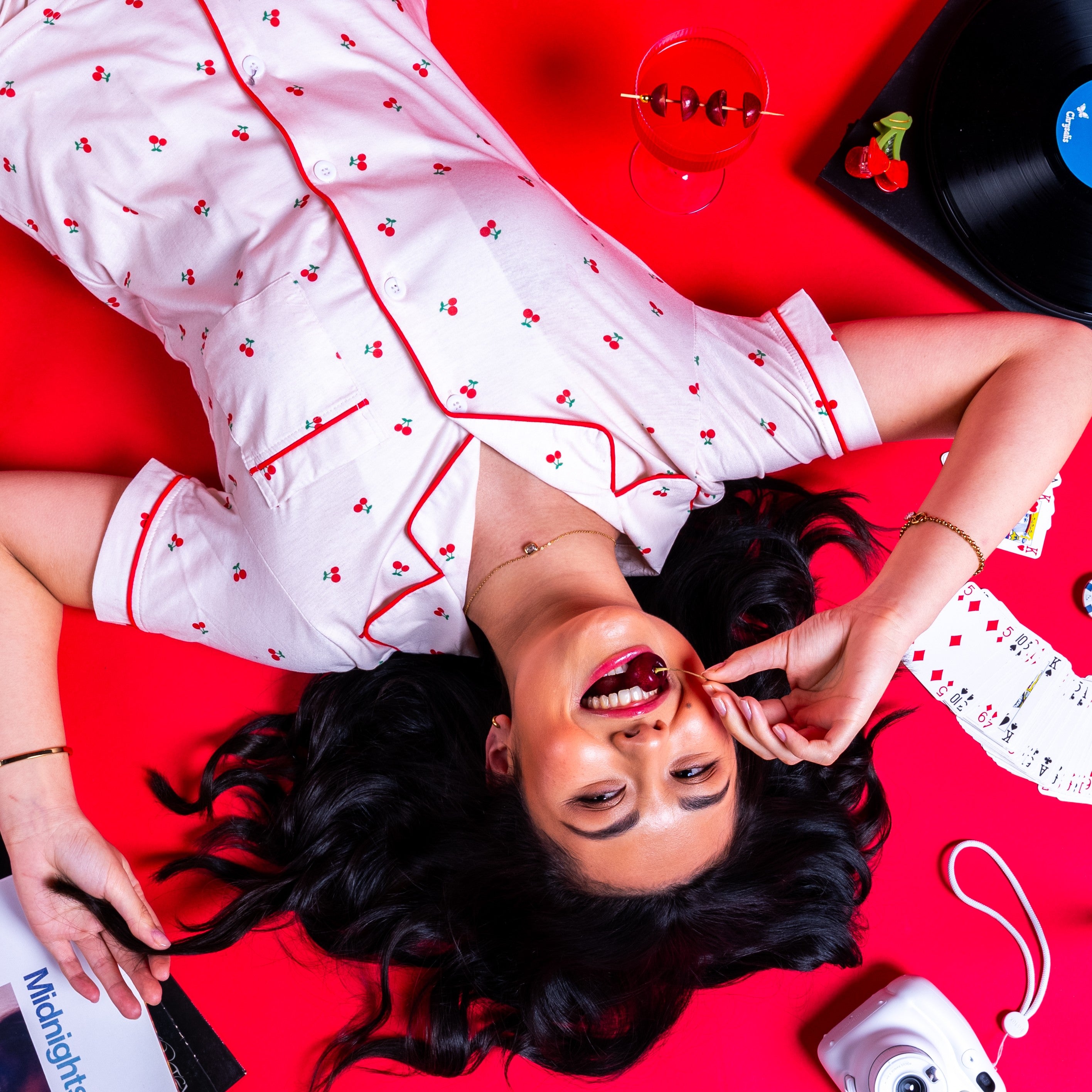 girl lying on the floor with cherry in mouth and cocktail, with retro background of camera, vinyl and cards