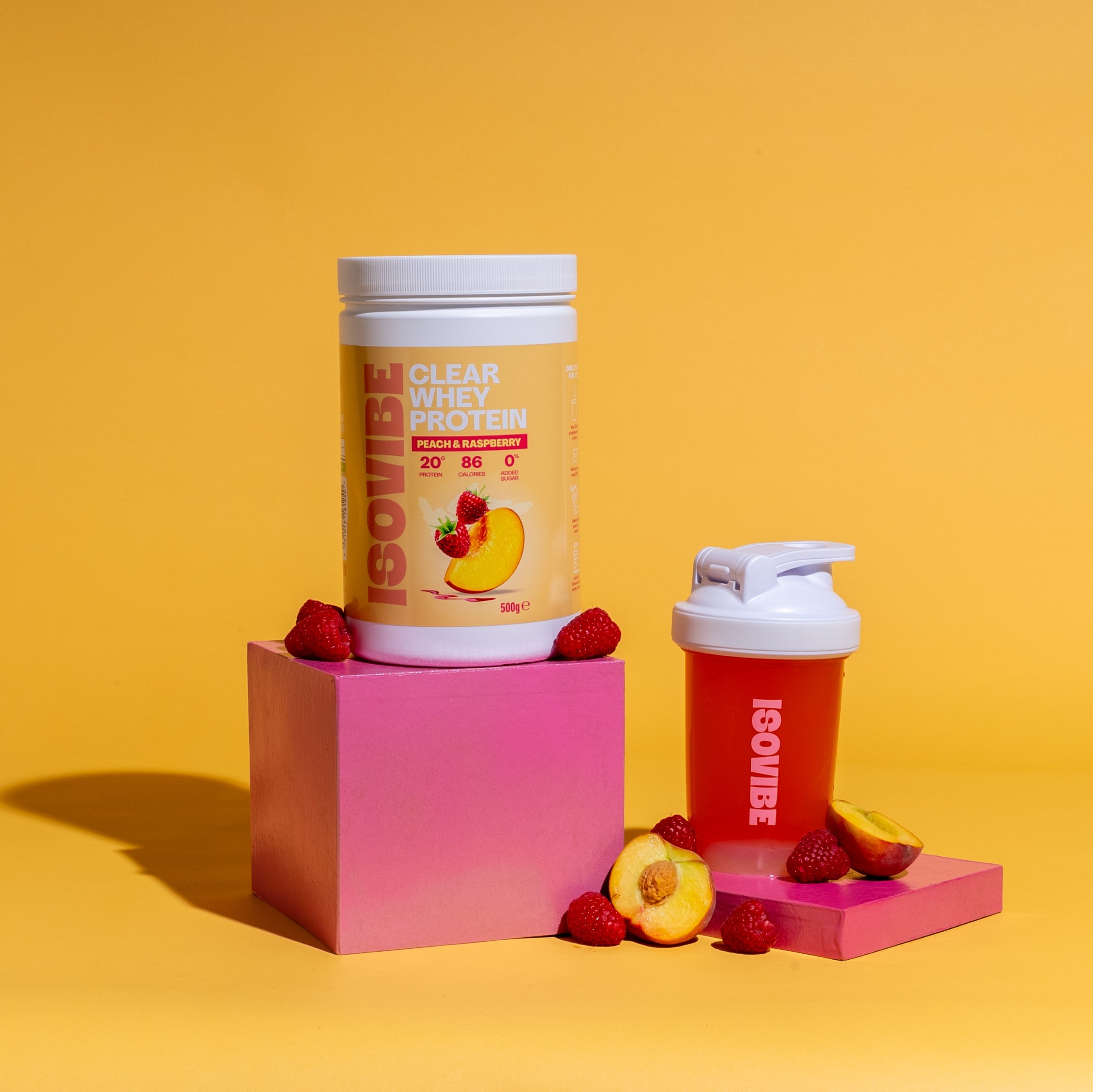 ISOVIBE Peach and Raspberry in a shaker and tub on podiums with sliced peaches and raspberries scattered around