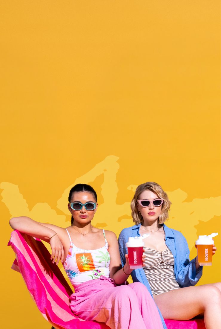 Two girls with sunglasses sat on a sun lounger holding drinks on a bright yellow background