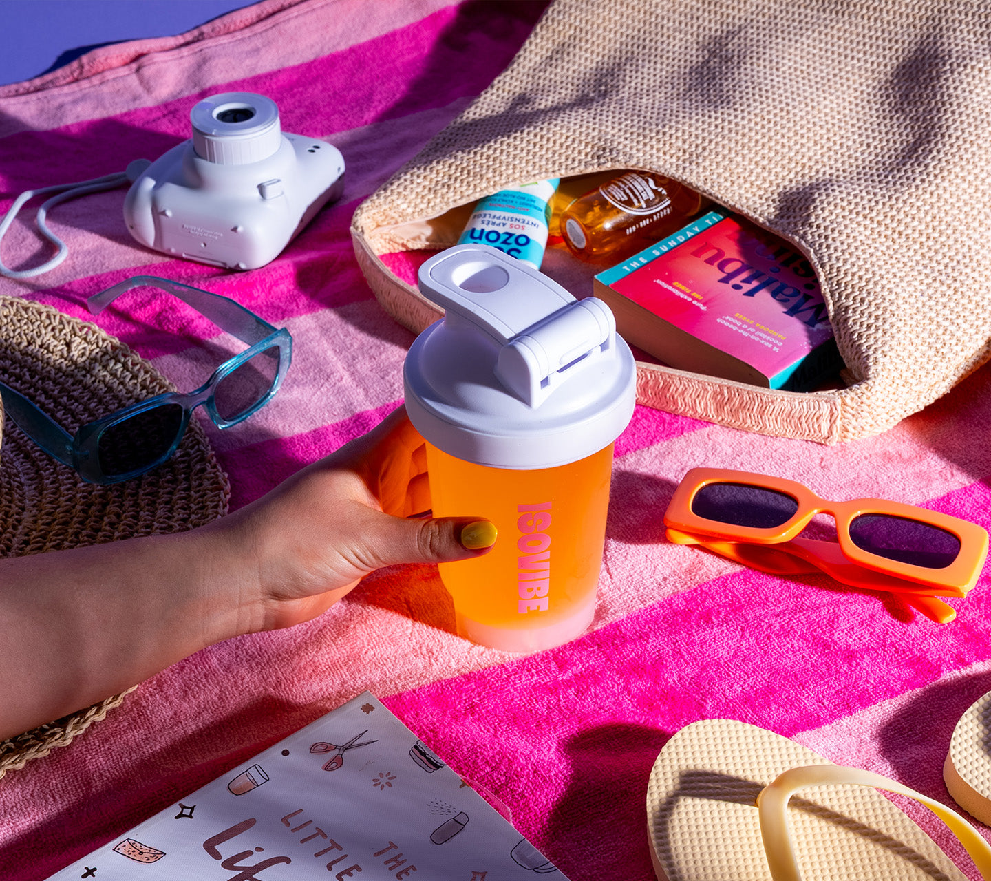 tropical isovibe in protein shaker alongside beach accessories, sunglasses, book and polaroid camera